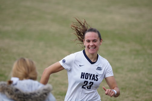 A female soccer player wearing a white jersey with 'Hoyas' and the number 23 printed on it is captured mid-motion, smiling with hair flying back. She appears to be on a grass field, engaging with someone in the foreground who wears a jacket with a fur collar.