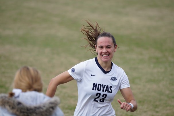 A female soccer player wearing a white jersey with 'Hoyas' and the number 23 printed on it is captured mid-motion, smiling with hair flying back. She appears to be on a grass field, engaging with someone in the foreground who wears a jacket with a fur collar.