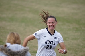 A female soccer player wearing a white jersey with 'Hoyas' and the number 23 printed on it is captured mid-motion, smiling with hair flying back. She appears to be on a grass field, engaging with someone in the foreground who wears a jacket with a fur collar.