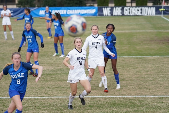 A soccer match is in progress with players wearing blue and white jerseys on a grassy field. Some players are actively engaged in the game, focusing on the ball which is suspended mid-air. Banners in the background indicate a championship setting, with the words 'NCAA Soccer Championship' and 'We Are Georgetown' visible.