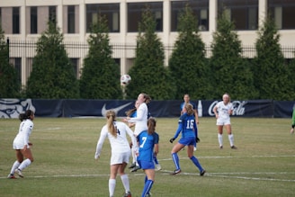 A group of female soccer players on a field are actively engaged in a game. Several players in blue and white uniforms are visible, with one player in the air heading the ball. In the background, there are trees, fencing, and a building.