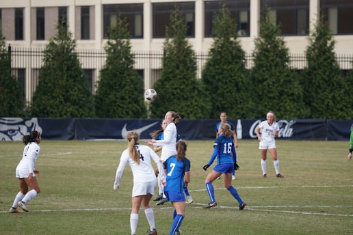 A group of female soccer players on a field are actively engaged in a game. Several players in blue and white uniforms are visible, with one player in the air heading the ball. In the background, there are trees, fencing, and a building.