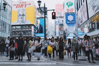 A busy urban scene featuring a crowd of people waiting at a crosswalk in an area surrounded by tall buildings adorned with large advertising billboards. Prominent advertisements include a beverage and various other products, creating a bustling city atmosphere.