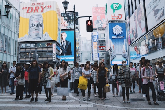 A busy urban scene featuring a crowd of people waiting at a crosswalk in an area surrounded by tall buildings adorned with large advertising billboards. Prominent advertisements include a beverage and various other products, creating a bustling city atmosphere.