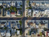 An aerial view of a residential neighborhood showing rows of houses with different colored roofs. The streets form a grid pattern, and there are numerous parked cars along the roads and driveways. The area is bordered with some greenery and trees, casting shadows on the ground.