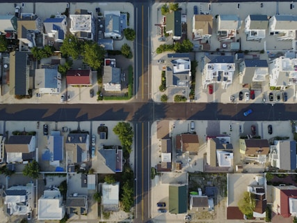 An aerial view of a residential neighborhood showing rows of houses with different colored roofs. The streets form a grid pattern, and there are numerous parked cars along the roads and driveways. The area is bordered with some greenery and trees, casting shadows on the ground.