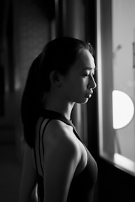A black and white photograph of a person in profile with a calm expression, standing near a window. The lighting creates a dramatic contrast, highlighting the subject's silhouette and facial features.