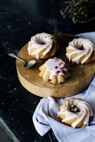Four small bundt cakes with white icing are arranged on a wooden round board. One of the cakes is decorated with red berries. A small silver spoon is placed on the board. A light blue napkin is draped underneath the board and one cake rests on it.