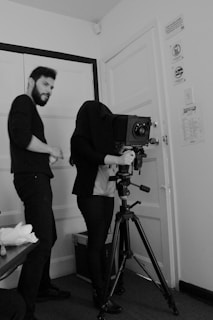 In a black and white room, a person is using a large vintage camera mounted on a tripod. Another person stands beside them, wearing dark clothing. The photographer has their head covered by a dark cloth, a technique used to focus the camera without light interference.