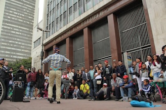 A street performer entertains a crowd of onlookers outside a large, official-looking building. The performer is dressed in casual attire, facing the audience with arms outstretched, possibly delivering a comedic act. The diverse crowd includes men, women, and children, some standing and others sitting or leaning against the building, which is marked as 'Banco de la República.' Nearby are musical equipment and props, suggesting a lively street performance.