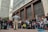 A street performer entertains a crowd of onlookers outside a large, official-looking building. The performer is dressed in casual attire, facing the audience with arms outstretched, possibly delivering a comedic act. The diverse crowd includes men, women, and children, some standing and others sitting or leaning against the building, which is marked as 'Banco de la República.' Nearby are musical equipment and props, suggesting a lively street performance.