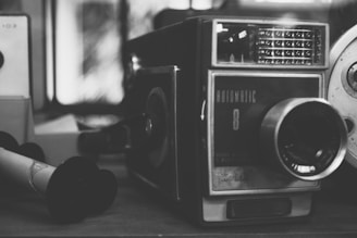 A vintage Kodak film camera with a prominent lens, detailed knobs, and labels. The camera appears to be resting among other indistinct objects in a softly lit environment, with a slightly blurred background.