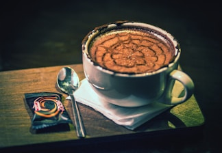 A cup of cappuccino with an intricate foam design sits on a wooden tray. Beside it, a small wrapped chocolate and a spoon are placed on a white napkin.