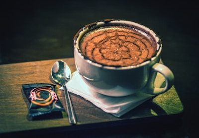 A cup of cappuccino with an intricate foam design sits on a wooden tray. Beside it, a small wrapped chocolate and a spoon are placed on a white napkin.