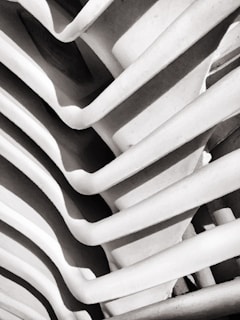 A close-up of a structural element featuring a series of curved, parallel lines or ribs creating a rhythmic pattern. The image is monochrome, highlighting the contrast between light and shadows.
