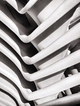 A close-up of a structural element featuring a series of curved, parallel lines or ribs creating a rhythmic pattern. The image is monochrome, highlighting the contrast between light and shadows.