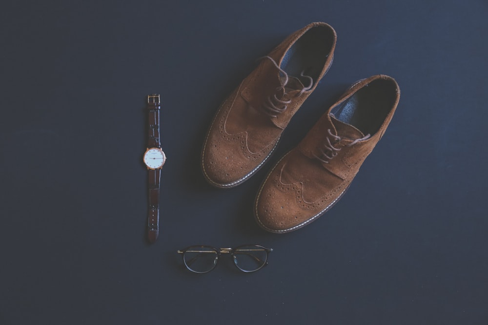 par de sapatos de ponta de asa Oxford de camurça marrom ao lado de óculos de armação preta e relógio analógico redondo dourado e branco com pulseira de couro marrom