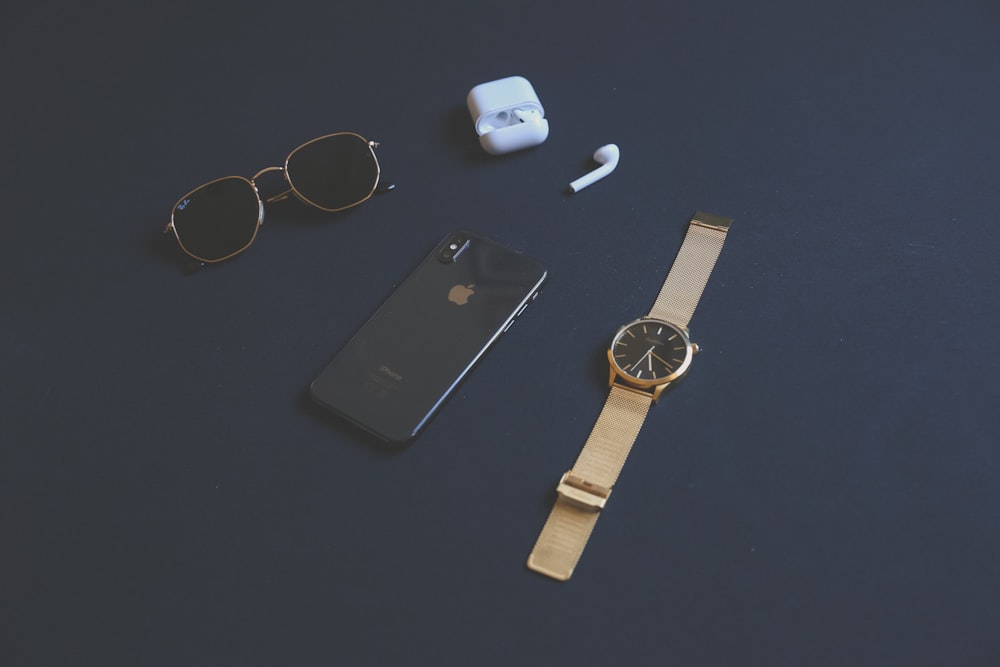 space gray iPhone X beside AirPods with case, round gold-colored analog watch and black sunglasses