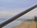 A metal handrail is prominently featured in sharp focus, diagonally traversing from the bottom left to the top right of the image. The background appears to be a blurry seaside view with a sandy beach on the right and some greenery in the distance. The sky has a gradient of light blue and soft pastel colors, suggesting a peaceful and calm atmosphere.