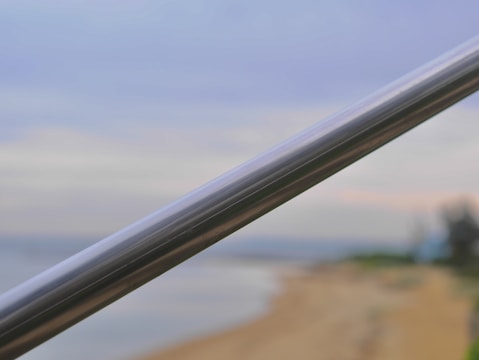 A metal handrail is prominently featured in sharp focus, diagonally traversing from the bottom left to the top right of the image. The background appears to be a blurry seaside view with a sandy beach on the right and some greenery in the distance. The sky has a gradient of light blue and soft pastel colors, suggesting a peaceful and calm atmosphere.
