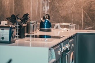 A modern kitchen counter featuring a variety of cooking appliances and utensils. A blue electric kettle and a metallic toaster are placed on the countertop alongside a stand holding cooking utensils. The design includes sleek cabinetry and a smooth, reflective surface with a neutral-toned backsplash.