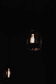 A dimly lit lantern with a visible glowing bulb inside, set against a dark background. The image conveys a minimalist and somewhat mysterious ambiance, with the focus on the warm light emanating from the lantern.
