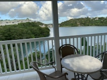 A balcony view overlooks a tranquil body of water surrounded by lush greenery and hills. There are several residential buildings visible in the distance. The foreground features a small round table and two chairs on a tiled floor, partially enclosed by a white railing. Fluffy clouds and patches of blue sky enhance the serene setting.