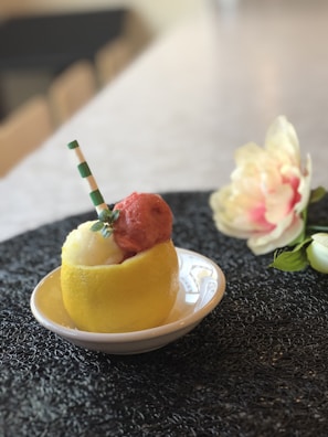 A creatively presented dessert with two scoops of sorbet, one red and one white, placed inside a hollowed-out lemon half. A striped straw stands vertically in the sorbet, and a small garnish is attached to it. The dish is positioned on a textured black placemat with a soft, blurred floral decoration in the background.