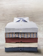 A neatly stacked set of five folded button-up shirts, each with different patterns and colors, arranged on a wooden surface.