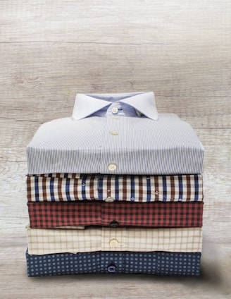 A neatly stacked set of five folded button-up shirts, each with different patterns and colors, arranged on a wooden surface.