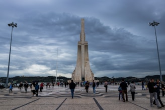 A large stone monument stands prominently against a cloudy sky, surrounded by a paved plaza with intricate patterns. Numerous people are walking around, suggesting a public or tourist area. Two tall streetlamps flank the scene, and hilly terrain is visible in the background.