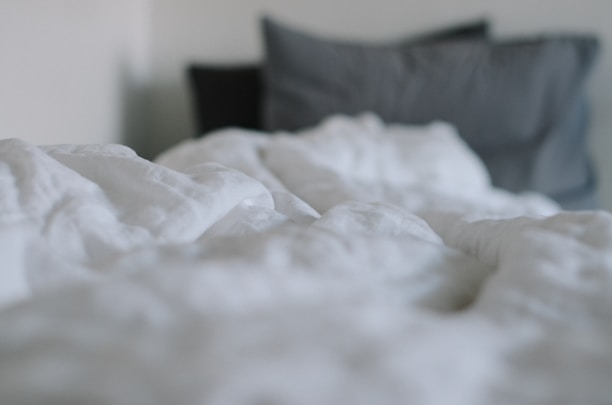 A soft, rumpled white blanket is in focus in the foreground, while two pillows, one dark gray and another slightly lighter gray, are in the background.