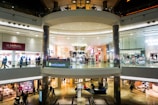 A spacious, multi-level shopping mall with various retail stores and a central open area. Shoppers walk along the corridors on both levels, surrounded by bright, well-lit storefronts displaying signs and advertisements. The architecture features large glass panels, modern light fixtures, and polished floors.