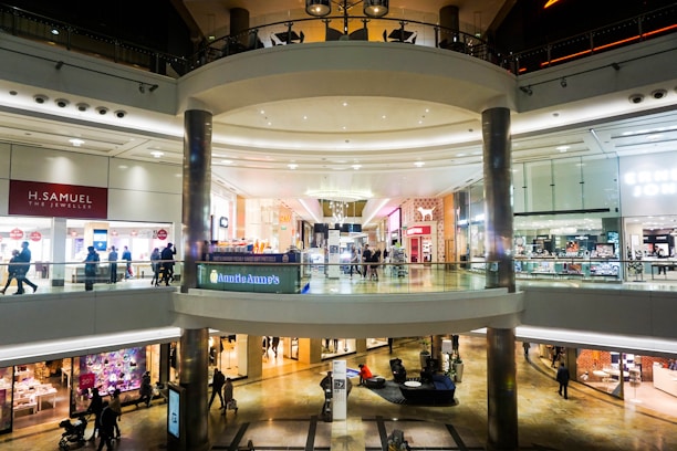 A spacious, multi-level shopping mall with various retail stores and a central open area. Shoppers walk along the corridors on both levels, surrounded by bright, well-lit storefronts displaying signs and advertisements. The architecture features large glass panels, modern light fixtures, and polished floors.