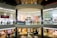 A spacious, multi-level shopping mall with various retail stores and a central open area. Shoppers walk along the corridors on both levels, surrounded by bright, well-lit storefronts displaying signs and advertisements. The architecture features large glass panels, modern light fixtures, and polished floors.