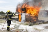 A firefighter wearing protective gear, including a helmet and breathing apparatus, is actively extinguishing a large fire engulfing a wooden structure. The flames are tall and intense, with thick smoke billowing upwards. The ground surrounding the structure is charred and there is foam visible, likely from the firefighting efforts.