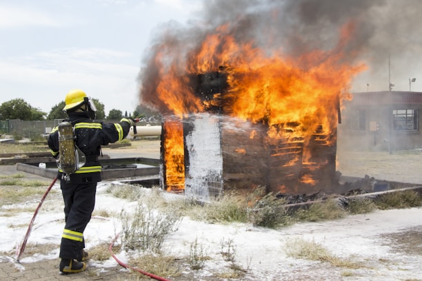 A firefighter wearing protective gear, including a helmet and breathing apparatus, is actively extinguishing a large fire engulfing a wooden structure. The flames are tall and intense, with thick smoke billowing upwards. The ground surrounding the structure is charred and there is foam visible, likely from the firefighting efforts.