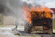 A firefighter is battling flames on a wooden structure using a hose, with water spray visible. Thick black smoke rises above the burning structure.