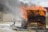 A firefighter is battling flames on a wooden structure using a hose, with water spray visible. Thick black smoke rises above the burning structure.