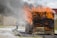 A firefighter is battling flames on a wooden structure using a hose, with water spray visible. Thick black smoke rises above the burning structure.
