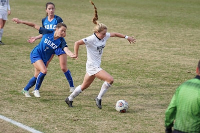 A soccer match between two teams, with one player in a white uniform actively dribbling the ball while two opposing players in blue are attempting to defend. The players are on a grass field, and a referee is visible wearing a green uniform.