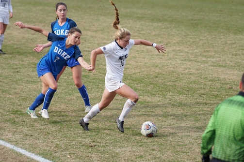 A soccer match between two teams, with one player in a white uniform actively dribbling the ball while two opposing players in blue are attempting to defend. The players are on a grass field, and a referee is visible wearing a green uniform.