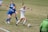 A soccer match between two teams, with one player in a white uniform actively dribbling the ball while two opposing players in blue are attempting to defend. The players are on a grass field, and a referee is visible wearing a green uniform.