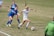 A soccer match between two teams, with one player in a white uniform actively dribbling the ball while two opposing players in blue are attempting to defend. The players are on a grass field, and a referee is visible wearing a green uniform.