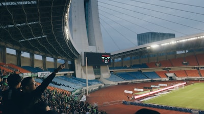 A large football stadium filled with empty orange and blue seats, with a crowd of spectators gathered in one section. The roof has a striking architectural design with cables and an overhanging structure, and the scoreboard displays a score of 2-1. The floodlights are turned on, suggesting an evening or late afternoon setting. A small number of players and officials are visible on the field near the goal.