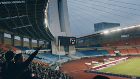 A large football stadium filled with empty orange and blue seats, with a crowd of spectators gathered in one section. The roof has a striking architectural design with cables and an overhanging structure, and the scoreboard displays a score of 2-1. The floodlights are turned on, suggesting an evening or late afternoon setting. A small number of players and officials are visible on the field near the goal.