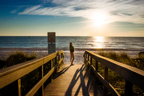 Golden hour light casting long shadows over a beachside boardwalk with footprints in the sand