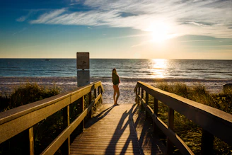 Golden hour light casting long shadows over a beachside boardwalk with footprints in the sand