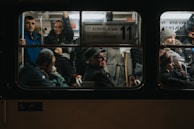 Inside a dimly lit public bus, a diverse group of passengers is visible through the window. People of various ages are engaged in different activities; some are seated, while others are standing. A man with a hat sits prominently in the center, appearing deep in thought, while nearby, passengers look outwards or converse. The atmosphere is crowded, indicating a busy commute.