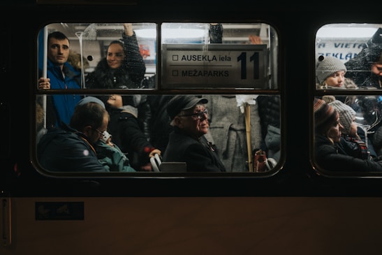 Inside a dimly lit public bus, a diverse group of passengers is visible through the window. People of various ages are engaged in different activities; some are seated, while others are standing. A man with a hat sits prominently in the center, appearing deep in thought, while nearby, passengers look outwards or converse. The atmosphere is crowded, indicating a busy commute.